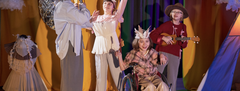 4 children on a stage with stars on their heads. One child is a wheelchair user.