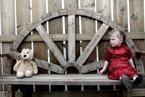 A young girl in a red dress sitting on the right of a long bench with a teddy at the opposite side of the bench.