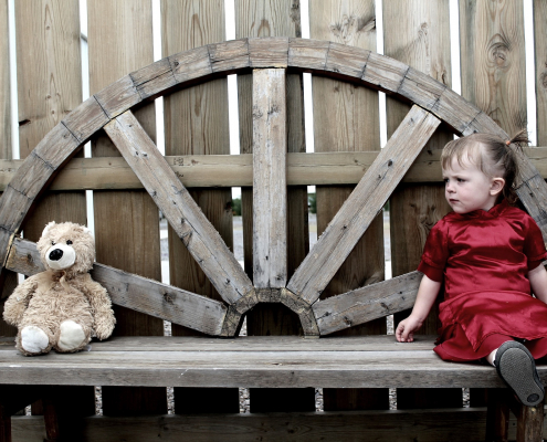 A young girl in a red dress sitting on the right of a long bench with a teddy at the opposite side of the bench.