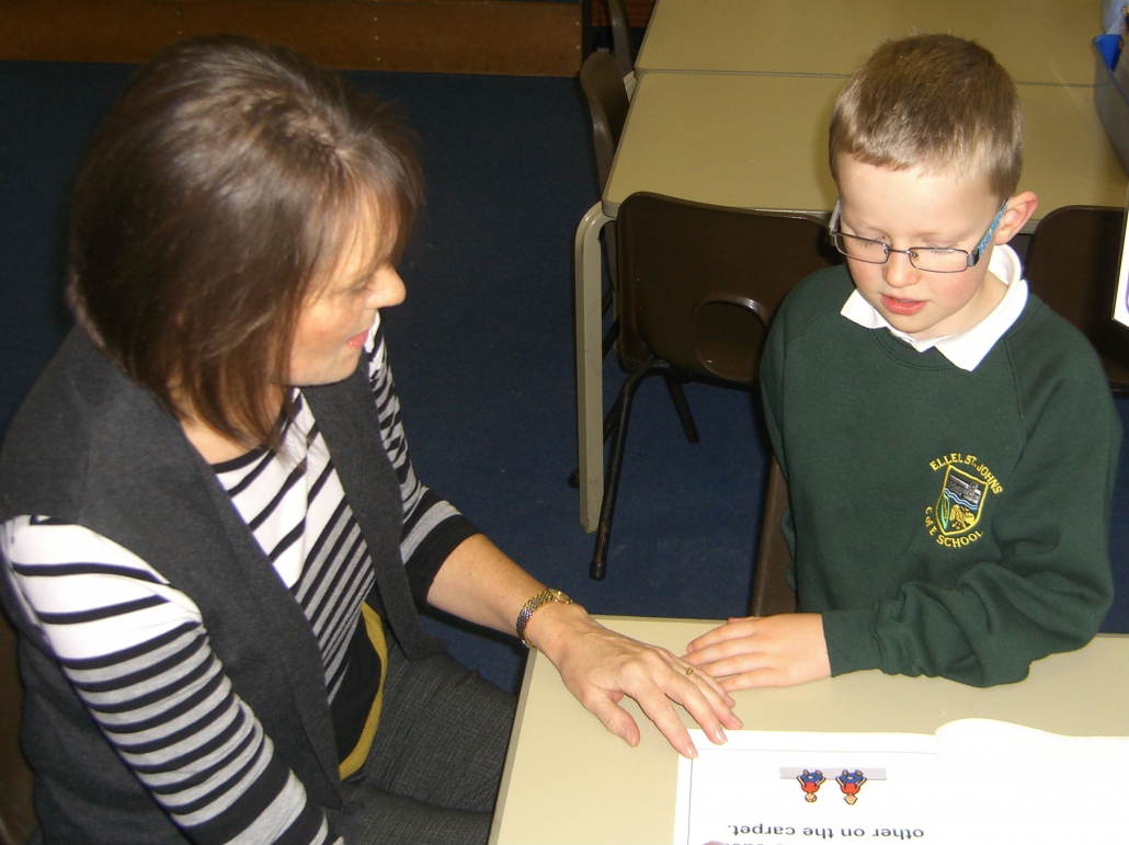 Teaching assistant sharing a story with a boy pupil in a green jumper. 