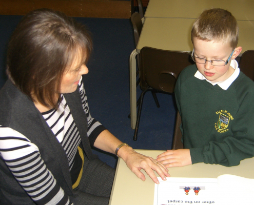 Teaching assistant sharing a story with a boy pupil in a green jumper.
