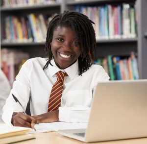 Black girl wearing a school uniform, sitting in a library working at a laptop.