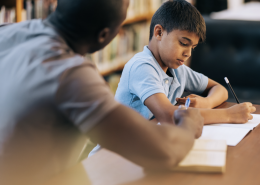 A teacher sitting at the side of a pupil helping them wit their work.