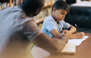 A teacher sitting at the side of a pupil helping them wit their work. 
