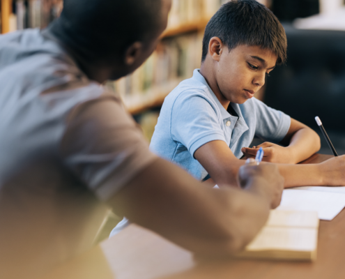 A teacher sitting at the side of a pupil helping them wit their work.