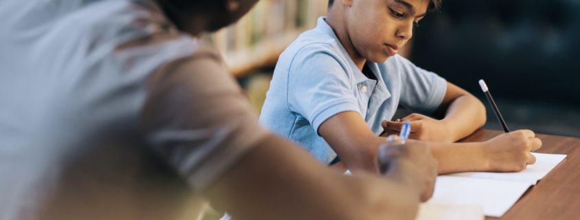 A teacher sitting at the side of a pupil helping them wit their work.