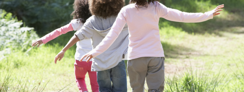 three young children following each other down a grassy path. You only seethe back of them.